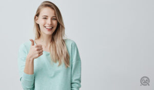 Happy young caucasian female wearing blue long sleeved shirt making thumb up sign and smiling cheerfully, showing her support and respect to someone